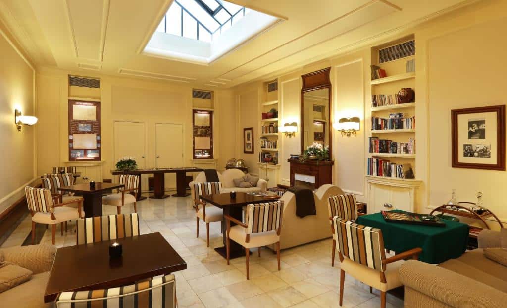 Sala de estar bem iluminada com cadeiras às riscas, mesas, estantes, sofás e uma clarabóia no teto, no Hotel Britania.