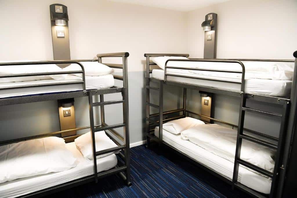 Quarto partilhado do YHA London St Pancras com dois beliches de ferro com almofadas e mantas, uma alcatifa azul no chão e candeeiros ao lado de cada cama.
