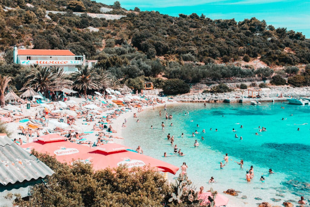 Praia de Pokonji Dol, com várias pessoas dentro do mar e uma faixa de areia branca à esquerda, com gente deitada em toalhas e sentada debaixo de chapéus de sol. Ao fundo, muita vegetação.