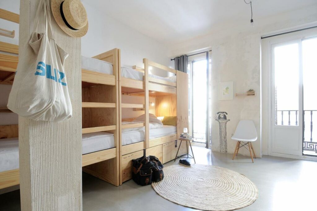 Quarto do The Hat Madrid. Beliches de madeira clara estão do lado esquerdo do quarto, com sacos e um chapéu pendurados. Há um tapete no centro do quarto e a parede do fundo tem duas grandes janelas com uma cadeira entre elas.