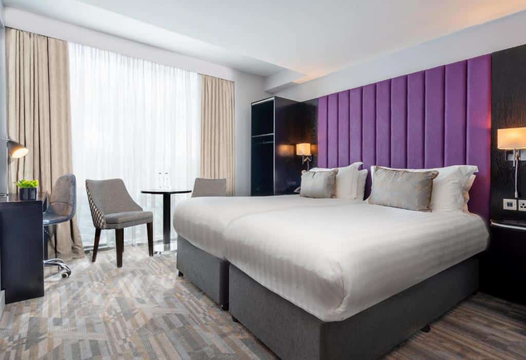 Quarto do The Trinity City Hotel com parede de cabeceira violeta, cama de casal branca, cadeira cinzenta e mobiliário escuro.