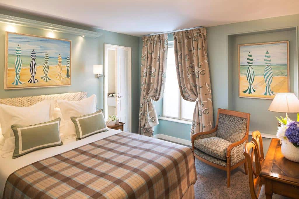 Quarto do Hôtel Le Relais des Halles com cama de roupa axadrezada, cadeira de madeira e janela com cortinas estampadas.