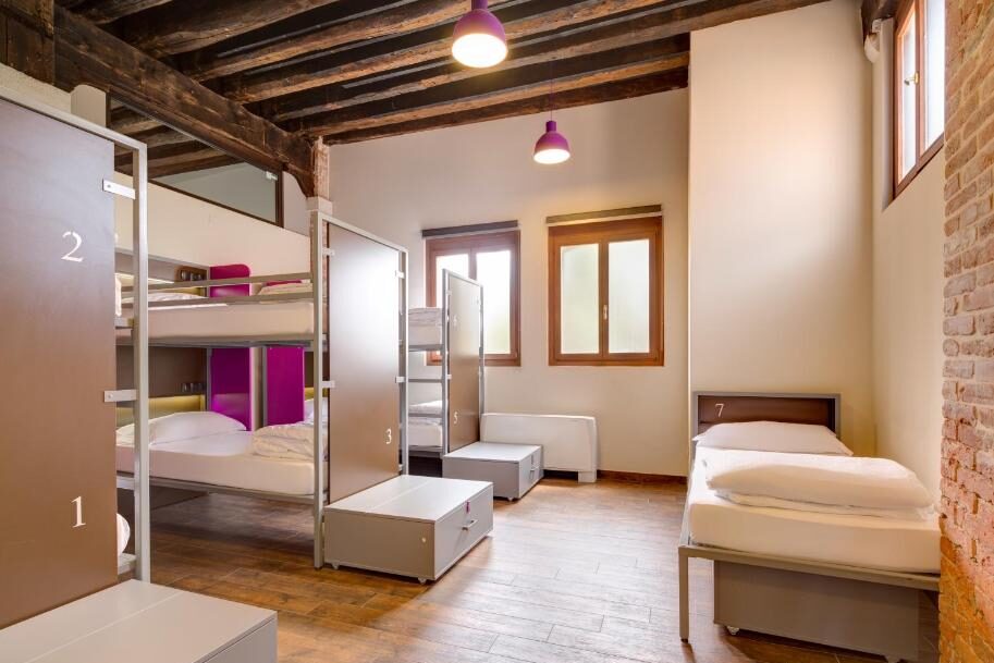 Quarto com beliche e cama de solteiro, paredes brancas, teto com vigas de madeira à vista e luzes LED cor-de-rosa escuro