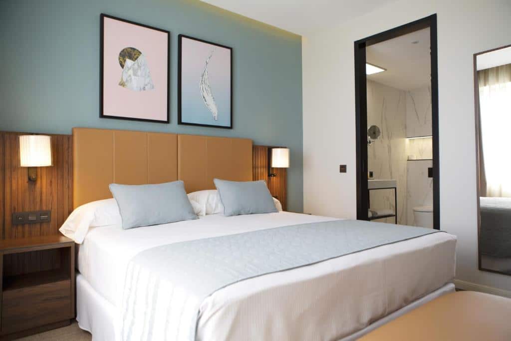 Quarto do Riu Plaza España, um dos melhores hotéis em Madrid. A cama grande tem roupa de cama branca e azul, e a cabeceira é acolchoada no centro e de madeira nas extremidades, com mesinha e candeeiro. A porta da casa de banho fica ao lado e está aberta, havendo também um espelho ao lado da porta. A parede da cama é azul e tem dois quadros.