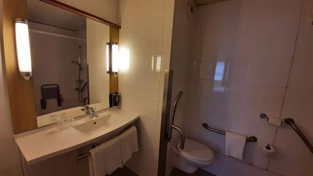Casa de banho adaptada do Ibis Lisboa José Malhoa com barras de apoio, espelho, lavatório mais baixo e cabine de duche sem vidro com cadeira