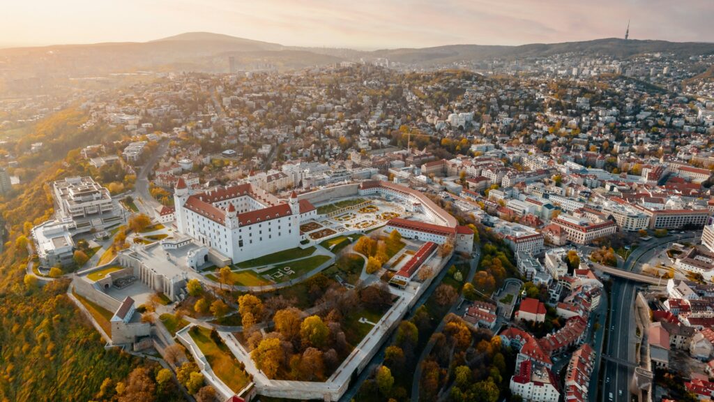 Vista aérea de Bratislava ao entardecer, com o Castelo de Bratislava em destaque no centro, rodeado por um vasto parque verde. O castelo tem uma arquitetura renascentista, com paredes brancas e telhados vermelhos. À volta do castelo, a cidade espalha-se com uma mistura de edifícios modernos e históricos em tons de vermelho e branco. Colinas suaves ao fundo e um céu suavemente iluminado pelo sol poente acrescentam uma atmosfera acolhedora à cena.