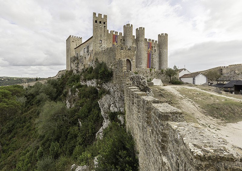 Castelo de Óbidos com várias torres, rodeado de vegetação sob um céu nublado, com um caminho a levar à entrada.