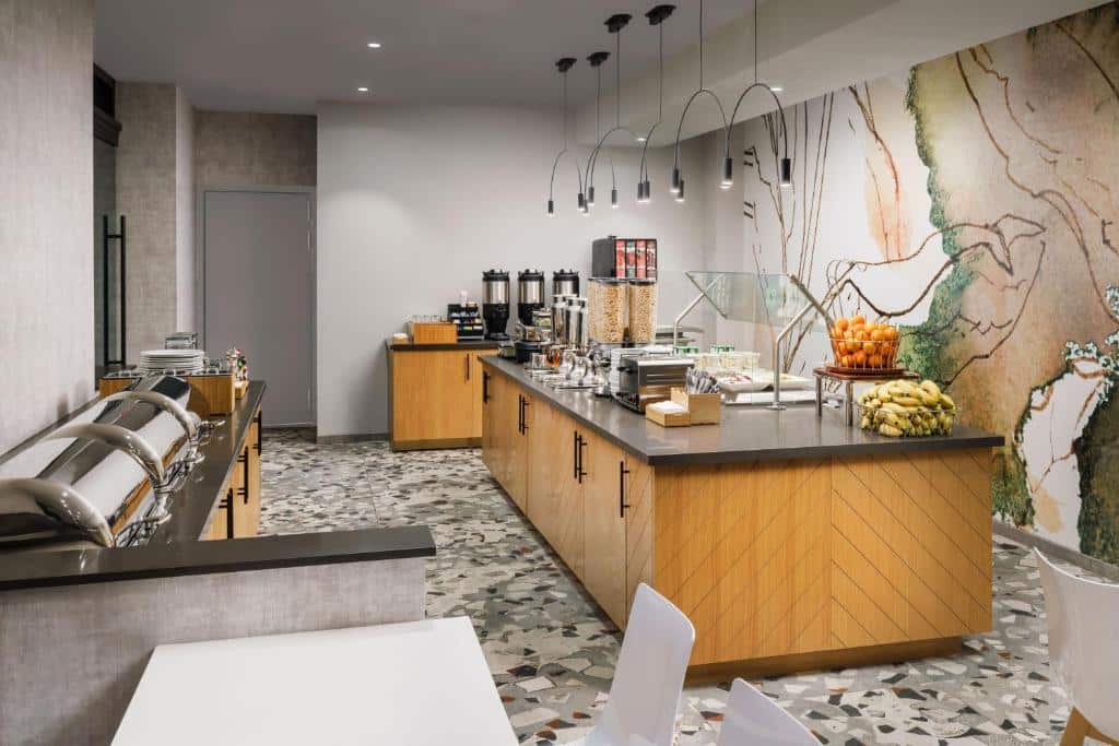 Zona de pequeno-almoço do SpringHill Suites com um buffet organizado, com fruta fresca, cereais, pão e bebidas. Decoração moderna, com bancadas de madeira clara e candeeiros suspensos.