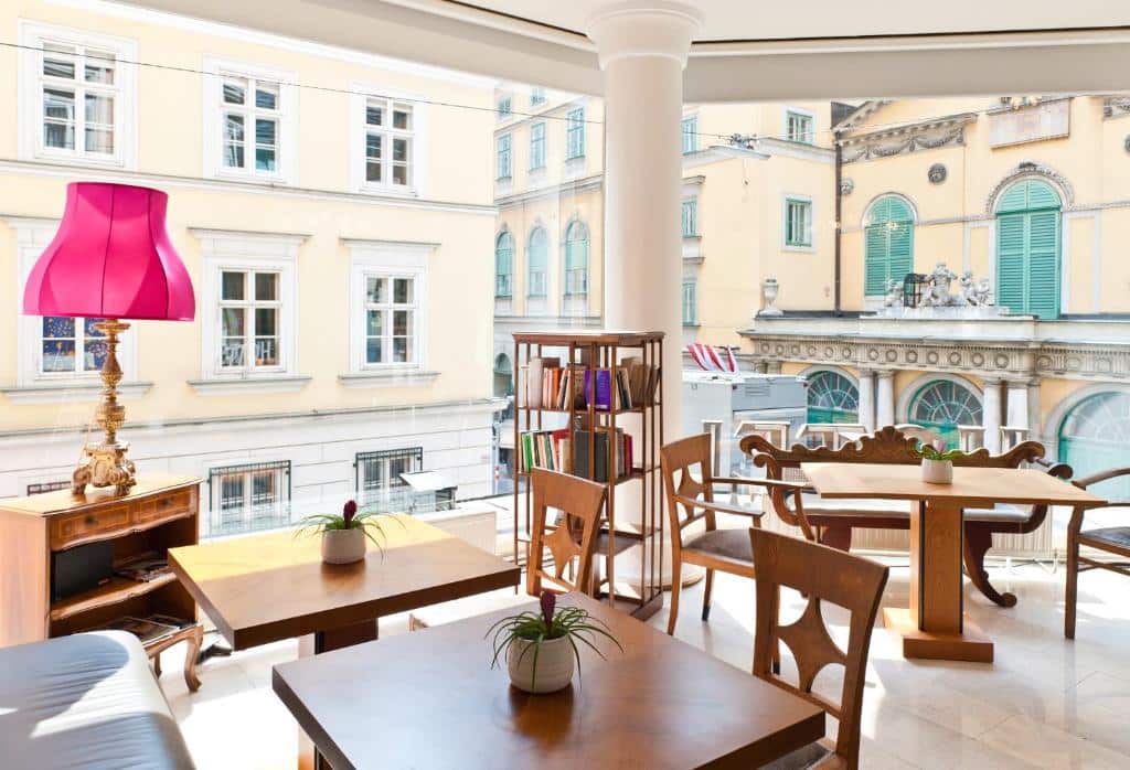 Café bem iluminado no Hotel Beethoven, com mesas e cadeiras de madeira, uma estante de livros e um candeeiro cor-de-rosa, com vista para os edifícios históricos.