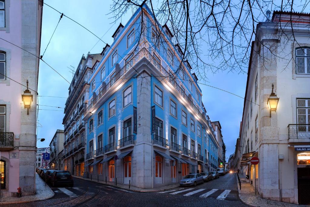 Edifício azul de esquina com varandas, onde fica o Lisboa Carmo Hotel, no cruzamento de ruas de calçada com candeeiros e carros estacionados.