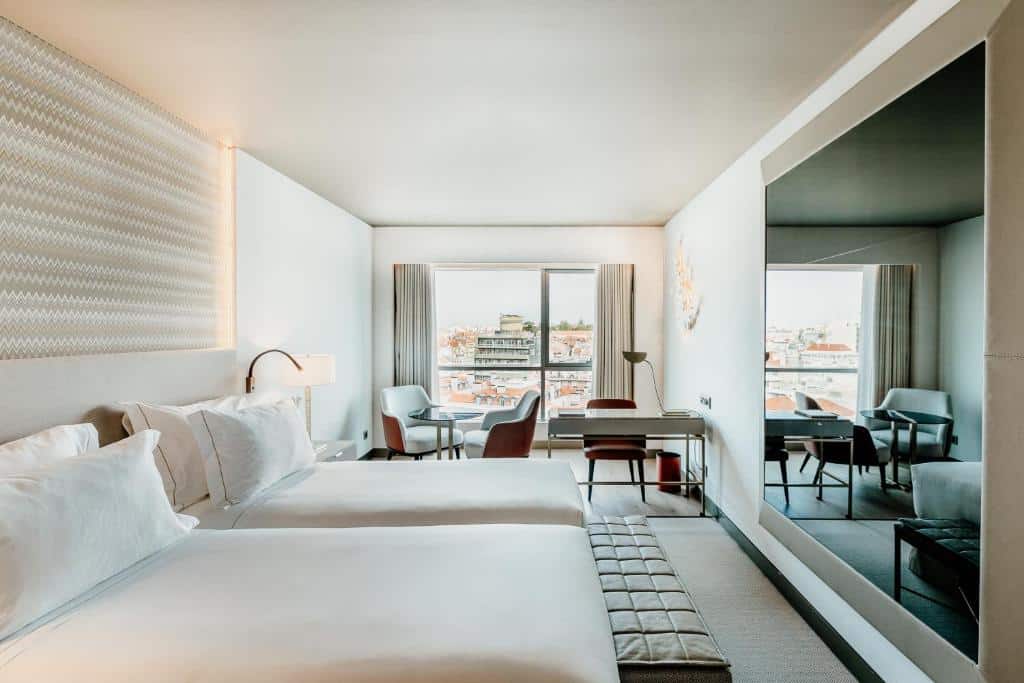 Quarto do EPIC SANA Marquês Hotel com duas camas individuais, uma varanda com cortinas e vista para a cidade, uma mesinha com poltronas junto à janela, uma secretária e um amplo espelho em frente às camas