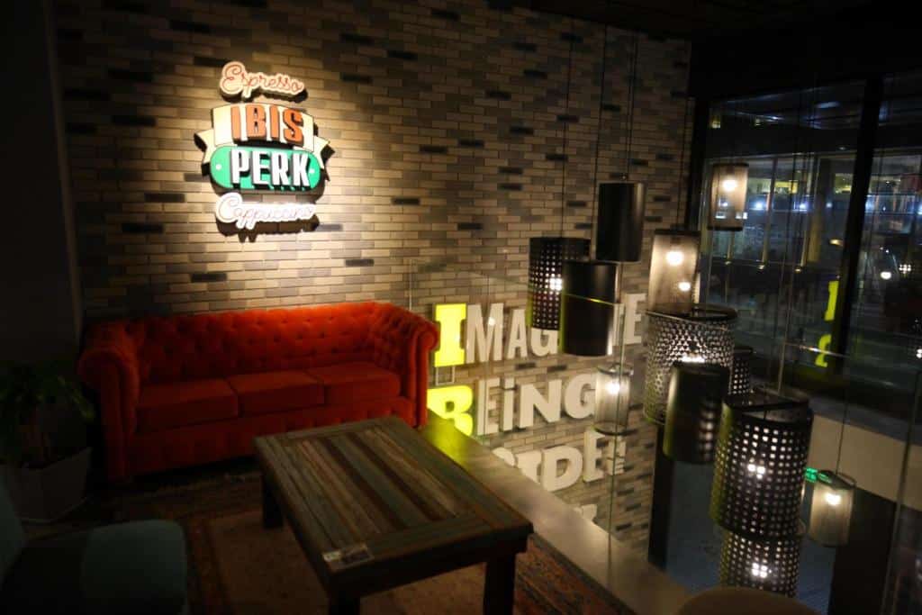 ambiente interior do ibis Styles Buenos Aires Florida, moderno, com um sofá vermelho de veludo junto a uma parede de tijolo à vista e uma placa iluminada, com atmosfera de café