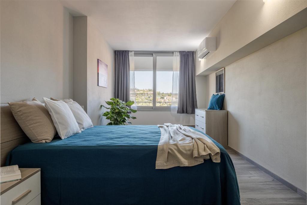 Quarto de casal moderno com cama azul, roupa de cama neutra e vista para zona urbana arborizada