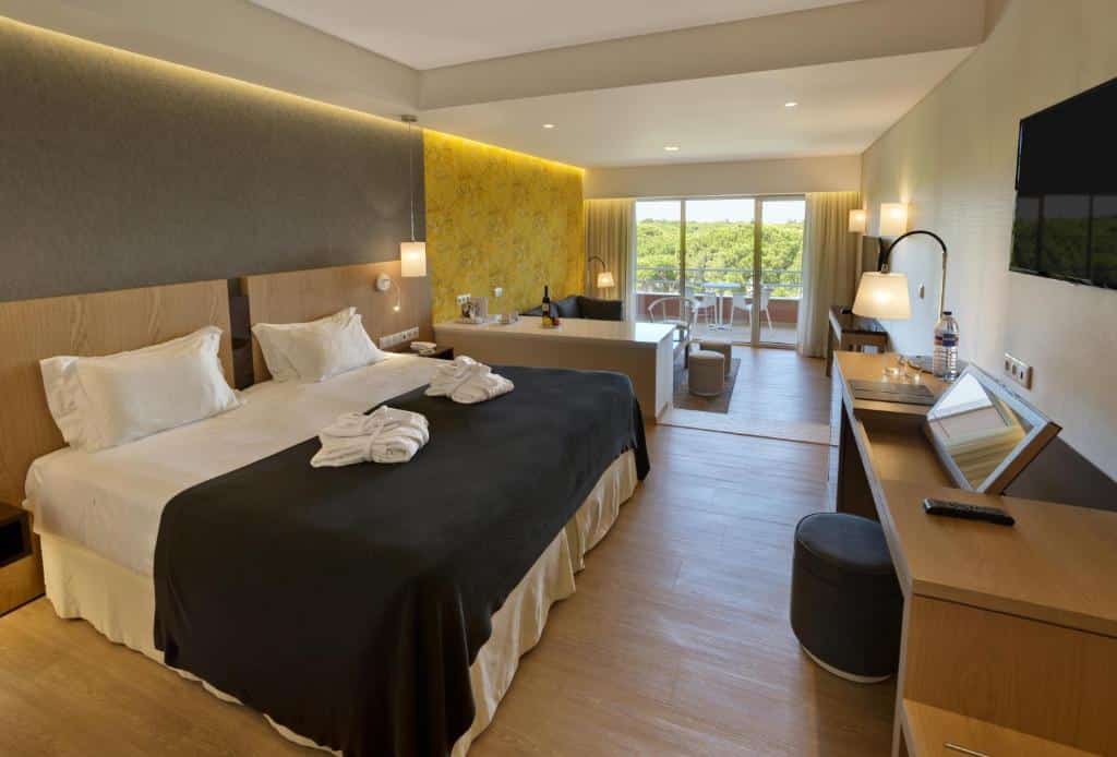 Quarto moderno no Falésia Hotel – Adults Only em Albufeira, com cama grande, secretária, TV, zona de estar e varanda com vista para a vegetação à luz do dia.