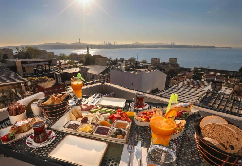Pequeno-almoço do Henna Hotel Istanbul servido numa mesa no terraço, com chá, sumo, pão e vários alimentos, com vista para a cidade e a água ao nascer do sol.