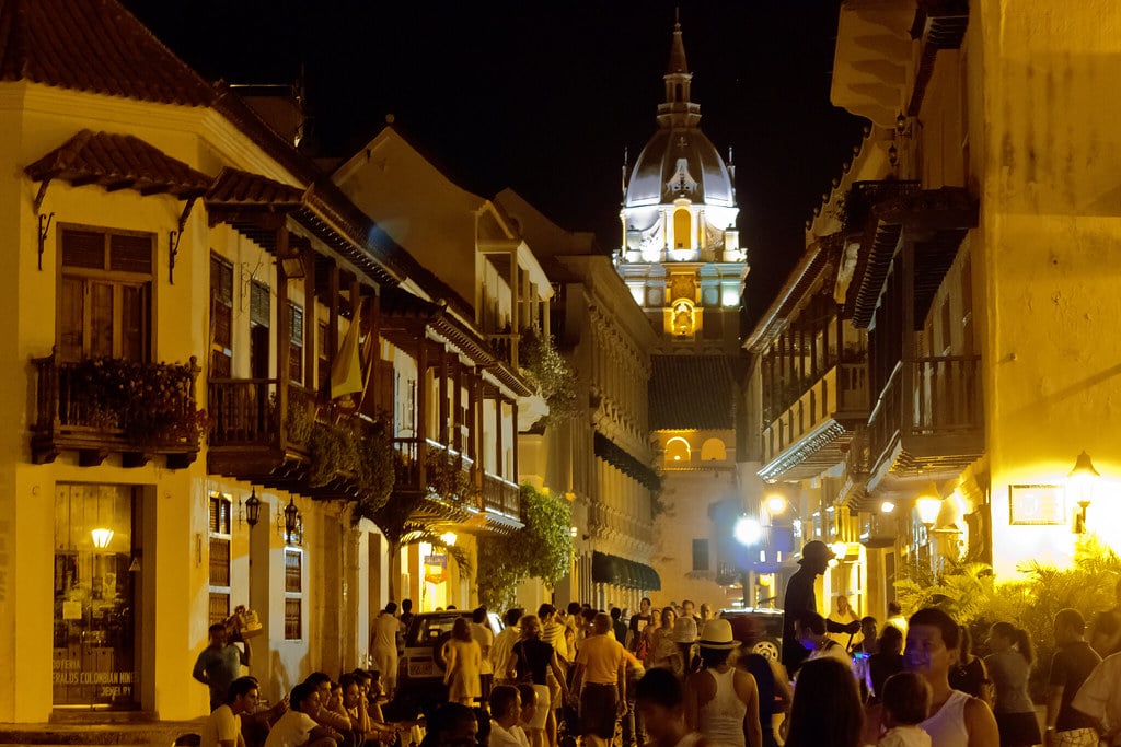 Rua movimentada ao fim do dia no centro histórico de Cartagena, com fachadas coloniais, varandas de madeira e uma torre iluminada ao fundo.