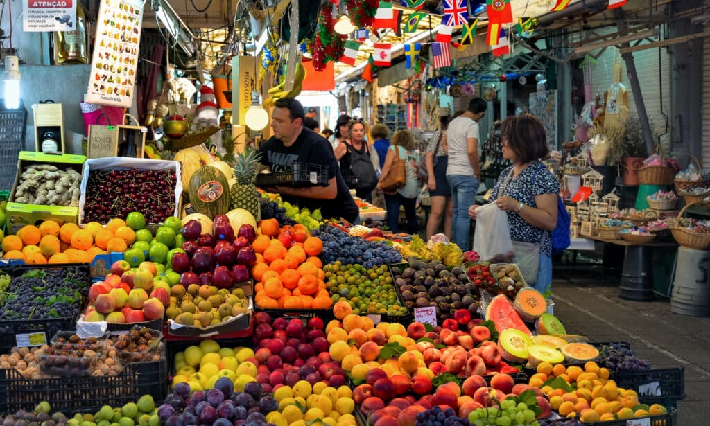 Interior do Mercado do Bolhão, com frutas expostas à esquerda e pessoas a passear ao fundo.