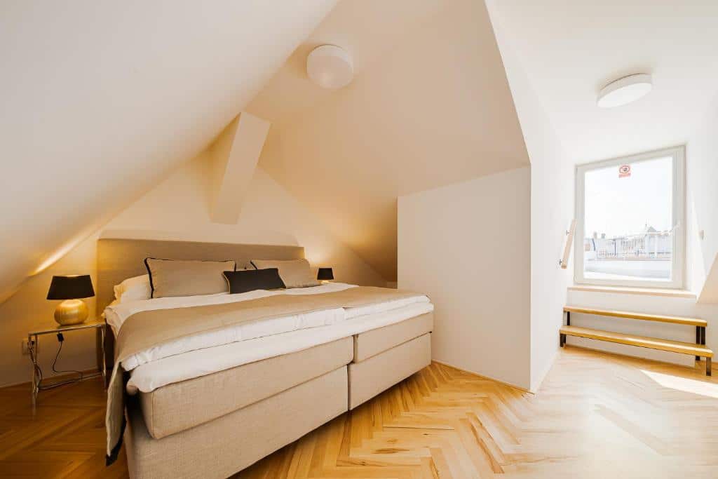 Quarto em águas-furtadas do Gold Art Apartments, com cama de casal, duas mesas de cabeceira com candeeiros e uma janela com degraus de madeira.