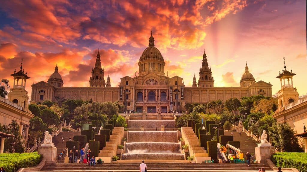 Palácio com uma fonte ao centro e escadarias nas laterais, com pessoas a passear em redor ao pôr do sol e o céu alaranjado.