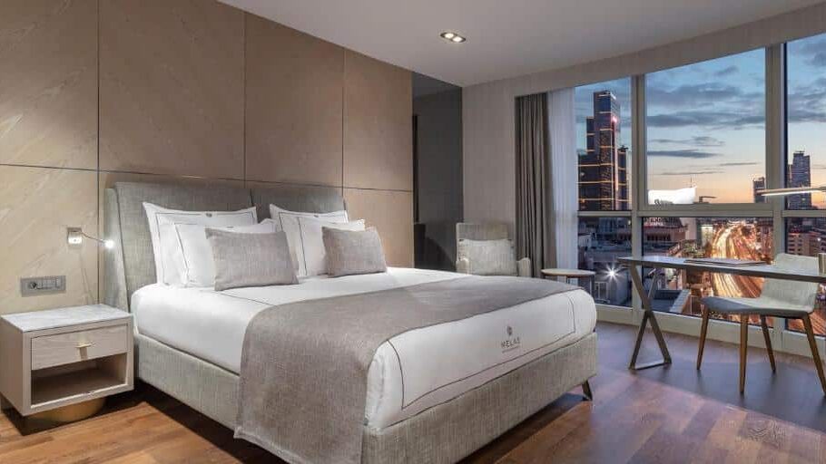 Quarto do Melas Hotel Istanbul com uma cama grande, mesa de cabeceira, secretária, cadeiras e vista da cidade através de uma grande janela ao pôr do sol.