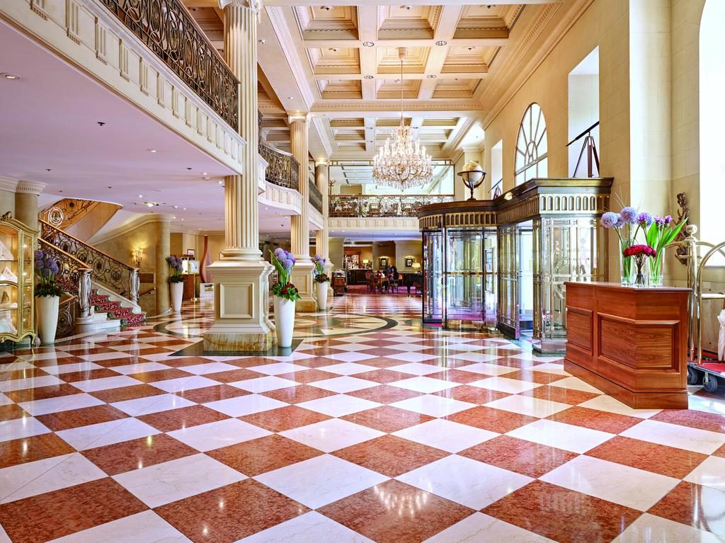Lobby elegante no Grand Hotel Wien, com chão de mármore axadrezado, colunas grandes, vitrinas de vidro, lustres e uma grande escadaria.