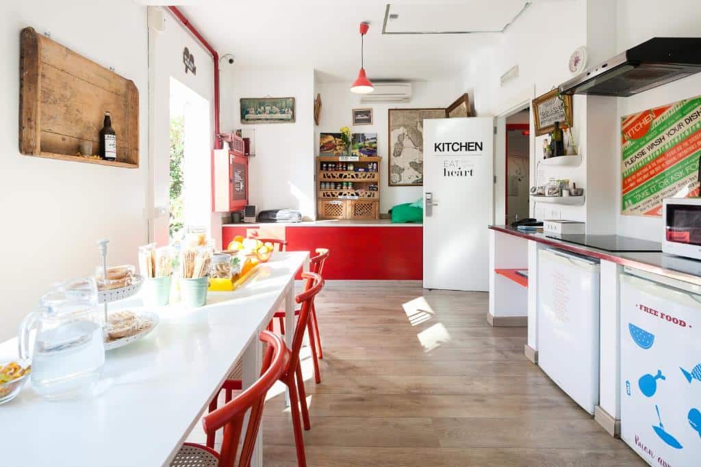 Cozinha partilhada do Ostello Bello com uma mesa com cadeiras vermelhas, uma bancada com eletrodomésticos e itens de pequeno-almoço sobre a mesa