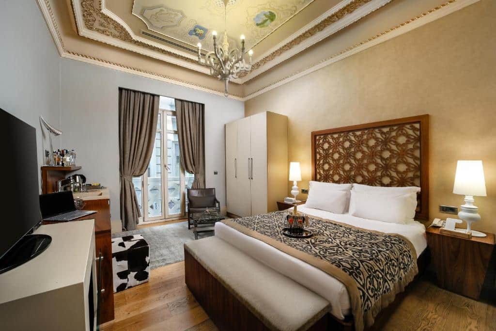 Quarto elegante do The Story Hotel Pera com uma cama de casal, teto ornamentado, lustre, TV, roupeiro e uma pequena varanda com cortinas.