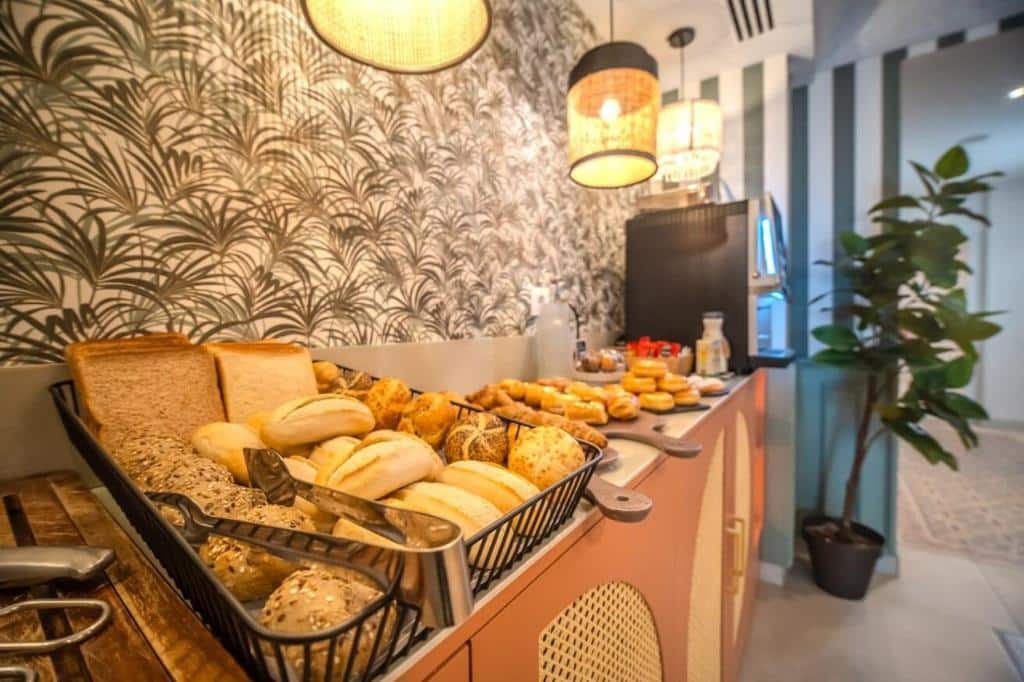 Pães e bolos variados num cesto de metal, numa cafetaria acolhedora do Soho Boutique Turia, com parede de estampa tropical e iluminação quente.