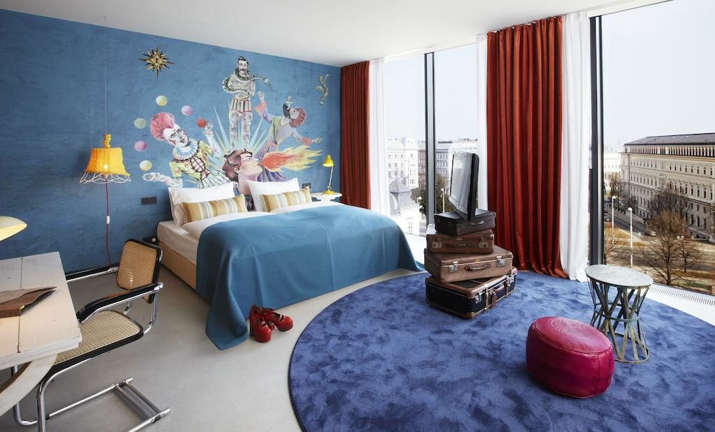 Quarto no 25hours Hotel, com tema azul, mural de circo, janelas grandes, malas empilhadas e mobiliário moderno.