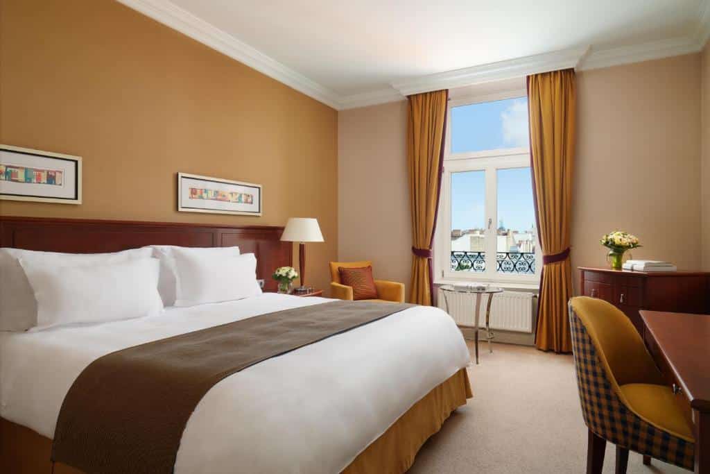 Quarto do Corinthia Budapest com uma cama grande, secretária, poltrona, janela com vista para a cidade e decoração em tons acastanhados.