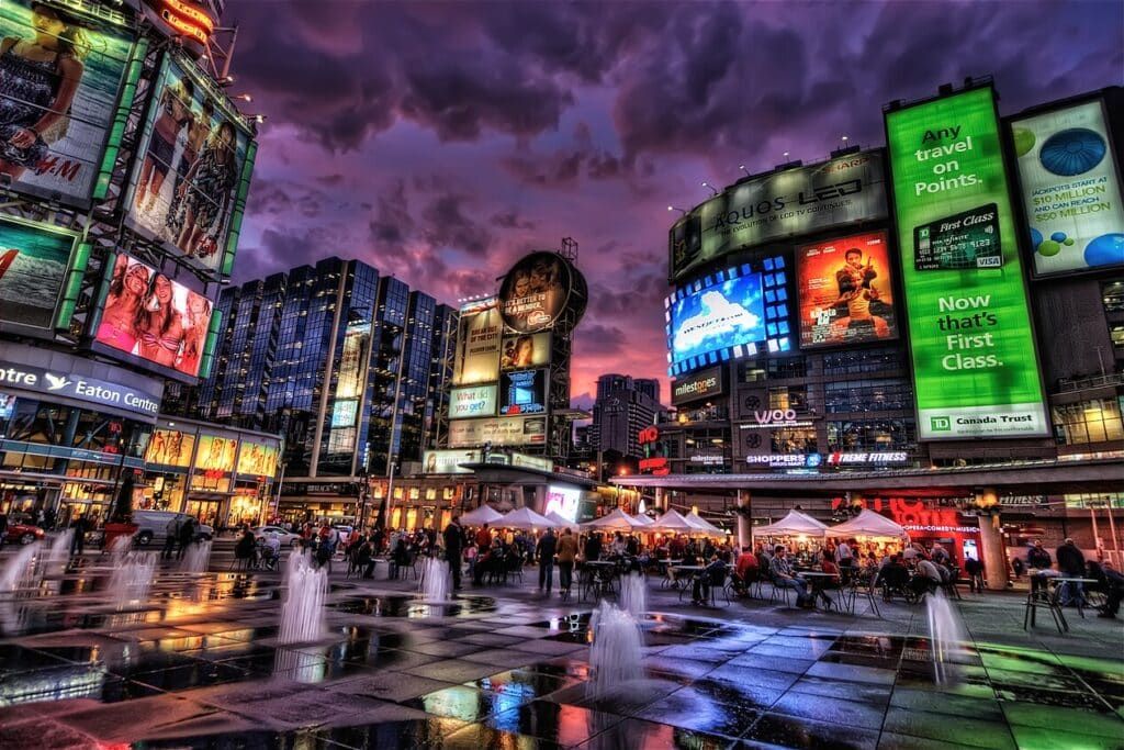 Centro de Toronto à noite, com edifícios iluminados, painéis publicitários e multidão numa praça com fontes em redor