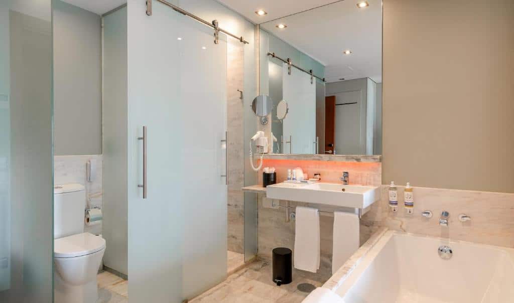 Casa de banho da Pousada de Lisboa - Small Luxury Hotels Of The World com banheira do lado direito, um lavatório com toalhas penduradas e um espelho amplo por cima, uma cabina de duche em vidro e uma sanita