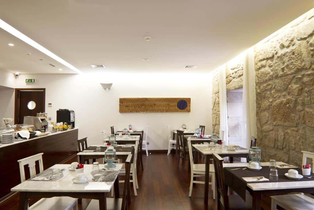 Pequena sala de refeições do Ribeira do Porto Hotel com várias mesas, chão de madeira, parede de pedra e um balcão com equipamento de café.