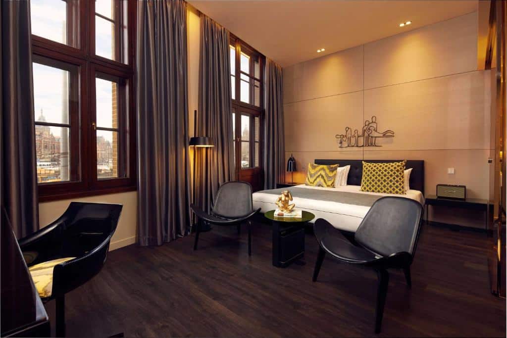 Quarto moderno do art’otel amsterdam, que ilustra onde ficar em Amesterdão: cama grande, cadeiras, pavimento de madeira escura, janelas altas e cortinados cinzentos.