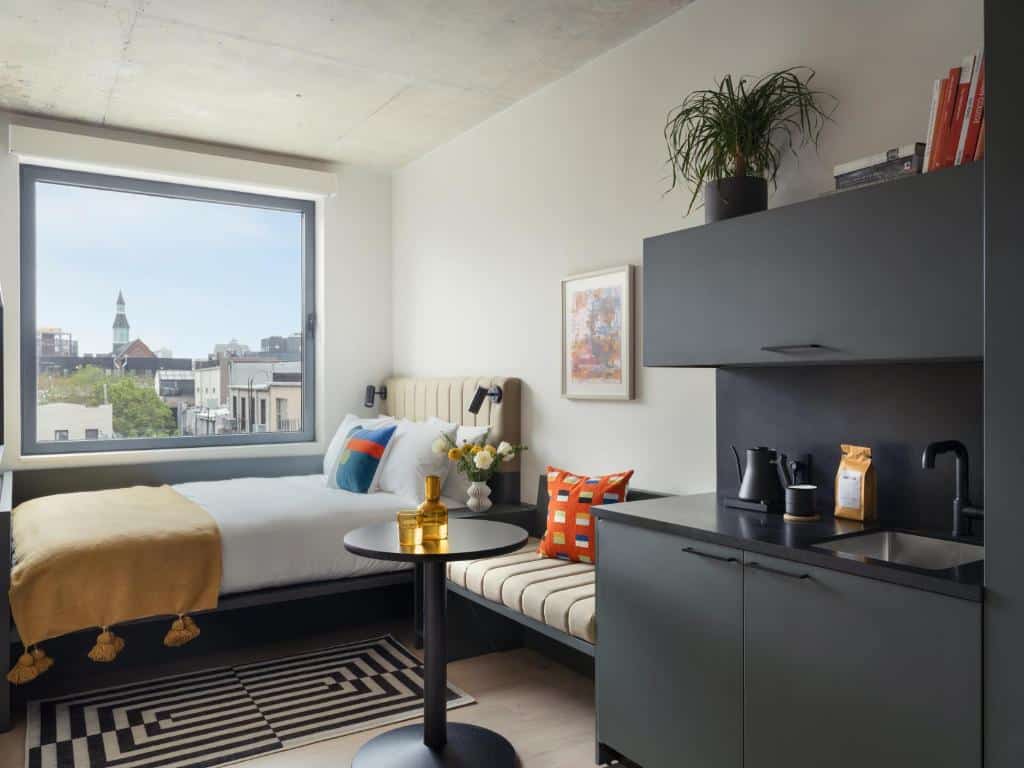 No quarto do Penny Williamsburg há uma cama de casal com colcha amarela e almofadas coloridas. À direita, uma pequena mesa redonda preta com um jarro dourado. Ao fundo, uma janela ampla com vista para a cidade; à esquerda, uma bancada preta com lavatório e máquina de café.