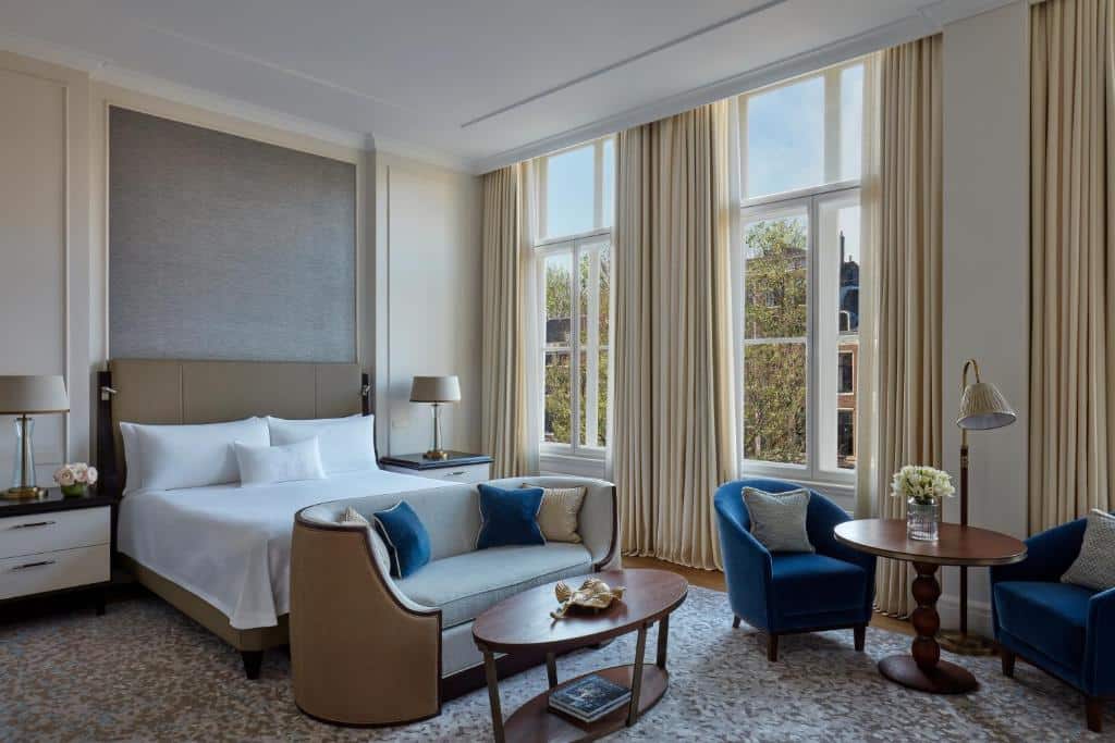 Quarto do Waldorf Astoria Amsterdam, que ilustra onde ficar em Amesterdão: cama king size, duas poltronas azuis, uma mesa redonda, janelas grandes com cortinados bege e decoração neutra.