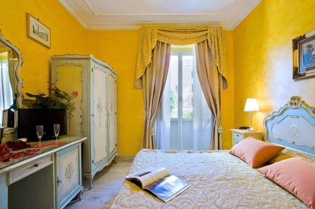 quarto do Hotel Villa San Lorenzo Maria com decoração clássica e requintada em tons de amarelo, cortinados pesados nas janelas altas, guarda-roupa grande a condizer com a cómoda em frente, televisão por cima e cama grande com mesa de cabeceira e candeeiro de ambos os lados