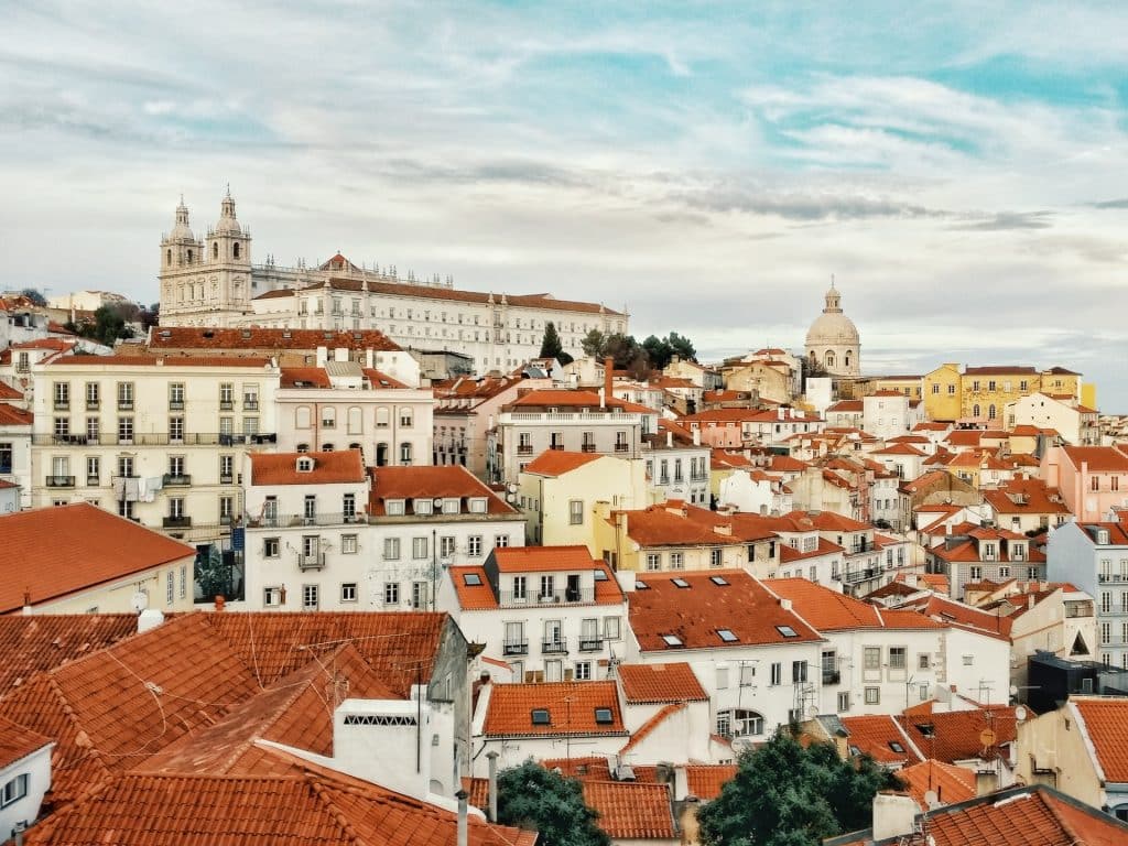 Conjunto de casas de telhado laranja a perder de vista pelas ruas de Lisboa, formando uma paisagem singular, com uma construção antiga ao fundo.