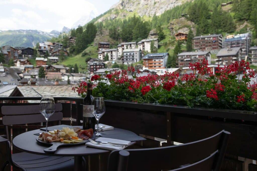 Varanda do Schweizerhof Zermatt, com mesa redonda, vinho e tábua de enchidos, com vista para a vila.