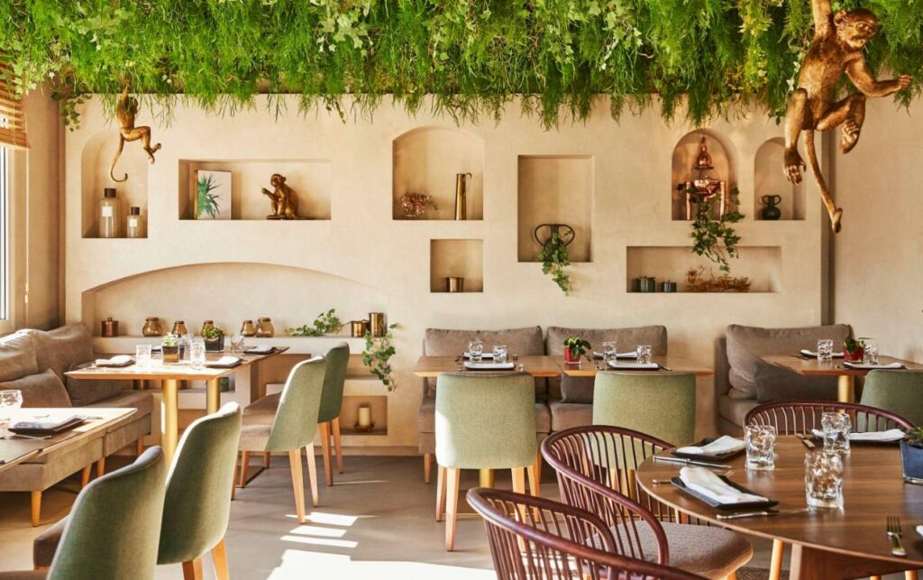 Zona de refeições do Hyatt Centric Gran Via Madrid decorada com o tema de selva. Mesas e cadeiras em tons castanhos e esverdeados estão dispostas na sala. O teto tem relva artificial e estátuas de macacos penduradas.