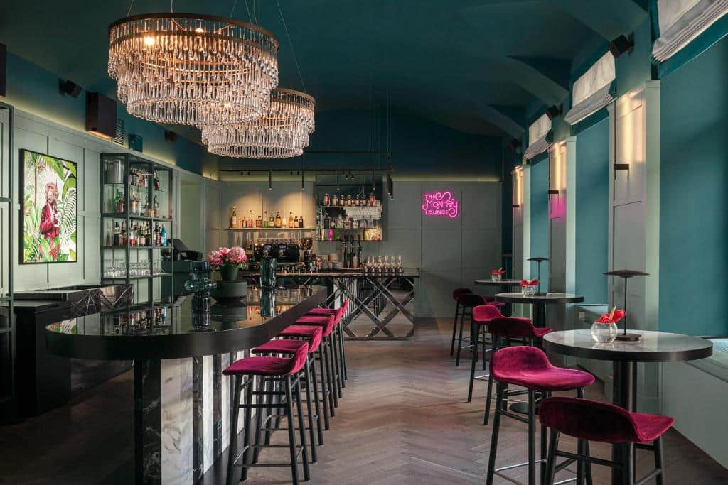 Bar moderno do Falkensteiner Boutique Hotel Prague, com paredes azul-petróleo, bancos de veludo vermelho e lustres de cristal.