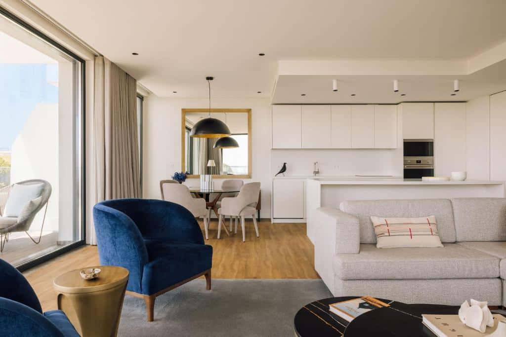 Cozinha compacta e sala de estar numa das unidades do Martinhal Lisbon Oriente, com um sofá e duas poltronas, uma varanda com cortinas e duas cadeiras estofadas no exterior, além de uma mesa com quatro lugares ao lado da cozinha