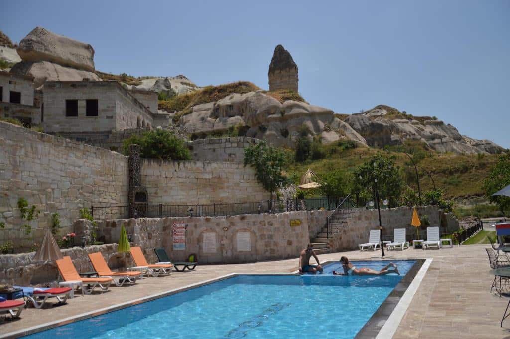 Duas pessoas a relaxar numa piscina exterior retangular rodeada de espreguiçadeiras, com colinas rochosas e edifícios de pedra ao fundo, no Grand Elite Cave Suites.
