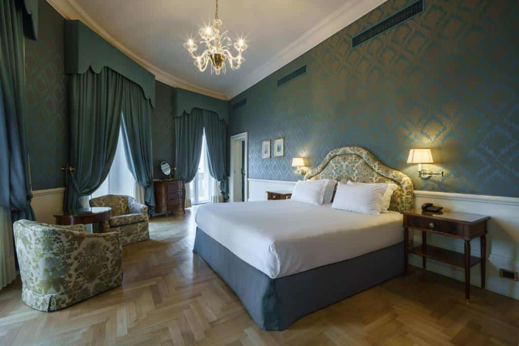 Quarto de luxo do Eurostars Hotel Excelsior com paredes de motivos florais azuis, cortinas verdes, cama de casal com dossel e mobiliário de madeira escura.