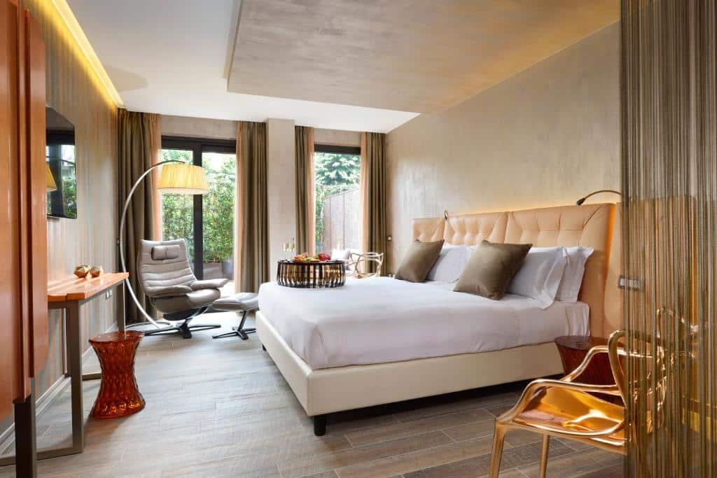Quarto do Milan Suite Hotel com varanda ampla ao nível do solo, cortinas, cama de casal espaçosa, poltrona com candeeiro de pé e televisão