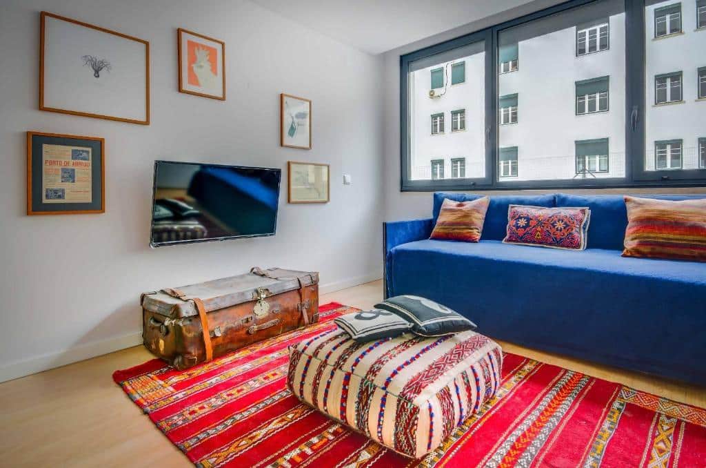 Sala de estar do Lisbon Serviced Apartments com sofá azul, TV na parede, arte emoldurada, baú de madeira, pufe estampado e tapete vermelho estampado.