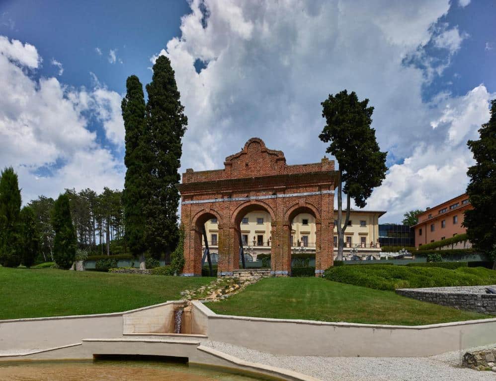 Entrada do Fonteverde Lifestyle &amp; Thermal Retreat, um dos hotéis na Toscana, com um portão alto de tijolo, muita vegetação — relvado verde baixo e árvores altas — e os edifícios do hotel atrás, um bege e outro laranja