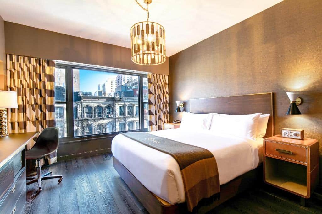 No quarto do Roxy Hotel há uma cama de casal com cabeceira de madeira à direita, com uma mesa de cabeceira com candeeiro e rádio. À esquerda, uma secretária com cadeira preta. Ao fundo, uma janela ampla com cortinados e vista para a cidade; no teto, um lustre suspenso.