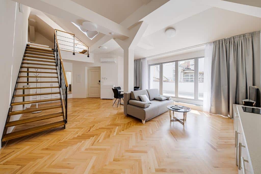 Apartamento loft moderno do Gold Art Apartments, com chão de madeira em espinha, sofá cinzento, escada, janelas grandes e zona de refeições.