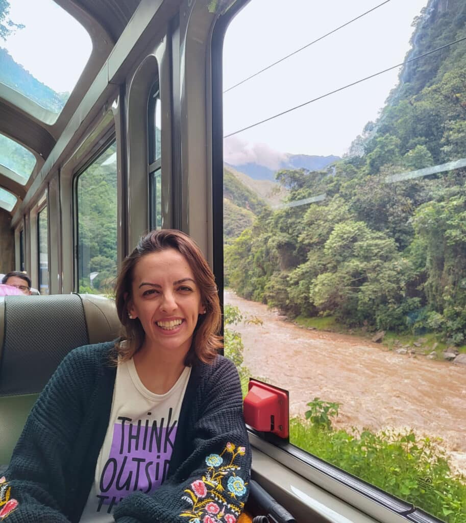 Passageira sentada junto à janela do vagão Vistadome da PeruRail, com as montanhas peruanas e o rio Urubamba lá fora